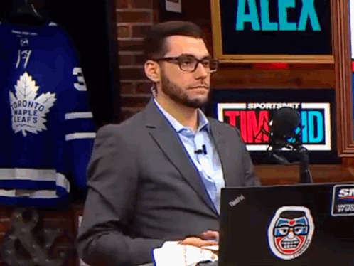 A Man In A Suit Is Sitting In Front Of A Laptop And A Sign That Says Alex On It GIF