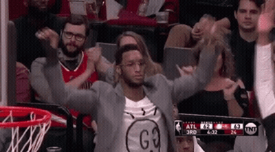 A Man In A Suit Is Sitting In The Stands At A Basketball Game With His Arms In The Air . GIF