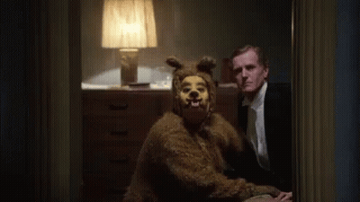 A Man In A Suit Is Sitting Next To A Bear Costume . GIF