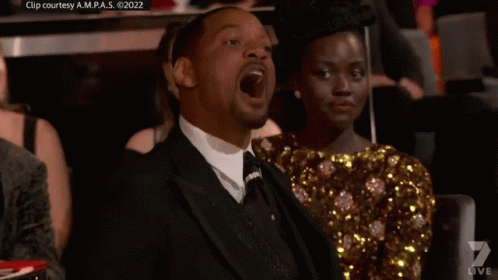 A Man In A Suit Is Sitting Next To A Woman In A Gold Dress GIF