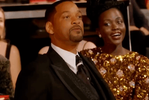 A Man In A Suit Is Sitting Next To A Woman In A Gold Dress . GIF