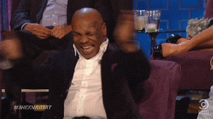 A Man In A Suit Is Sitting On A Couch With His Arms Outstretched And Laughing . GIF