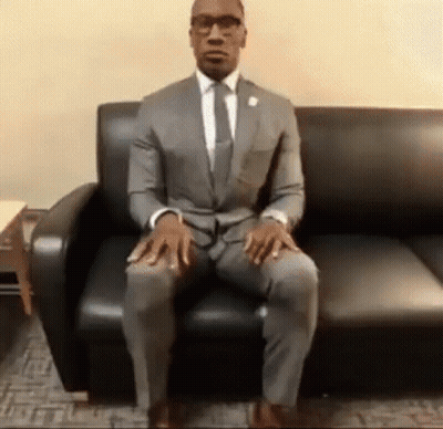 A Man In A Suit Is Sitting On A Couch With His Legs Crossed . GIF