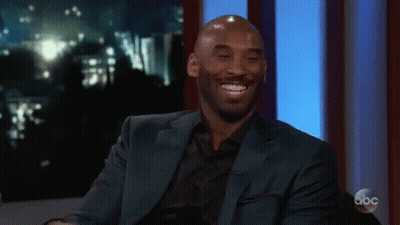 A Man In A Suit Is Smiling And Clapping His Hands While Sitting On A Stage . GIF