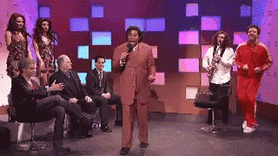 A Man In A Suit Is Speaking Into A Microphone While A Group Of People Sit Around Him GIF