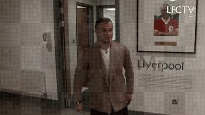 A Man In A Suit Is Standing In A Hallway Next To A Framed Picture Of A Man In A Red Shirt . GIF