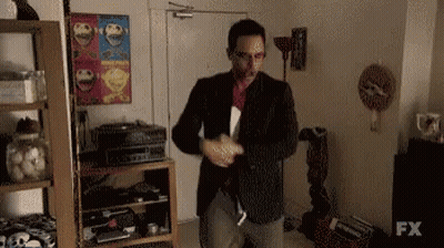 A Man In A Suit Is Standing In A Living Room With A Sign That Says Fx On It . GIF