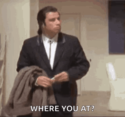A Man In A Suit Is Standing In A Room Holding A Jacket And Asking Where You Are . GIF