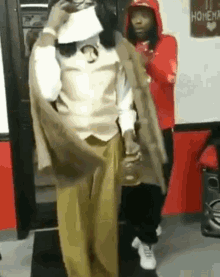 A Man In A Suit Is Standing In A Room With A Woman In A Red Hoodie Behind Him . GIF