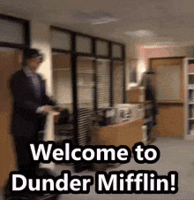 A Man In A Suit Is Standing In An Office With The Words Welcome To Dunder Mifflin Written On The Floor . GIF