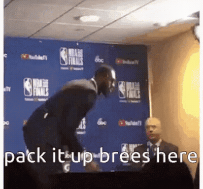 A Man In A Suit Is Standing In Front Of A Blue Wall With The Words Pack It Up Brees Here Written On It . GIF