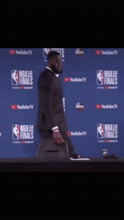 A Man In A Suit Is Standing In Front Of A Wall That Says Nba Finals On It GIF