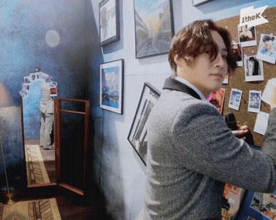 A Man In A Suit Is Standing In Front Of A Wall With Pictures On It And A Sign That Says Ithek GIF