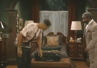 A Man In A Suit Is Standing Next To A Man In A Colorful Shirt Dancing In A Living Room GIF