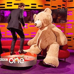 A Man In A Suit Is Standing Next To A Teddy Bear That Is Sitting On A Red Couch GIF