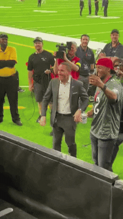 A Man In A Suit Is Standing On A Football Field Surrounded By People GIF