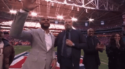 A Man In A Suit Is Standing On A Football Field With A Group Of People Applauding . GIF