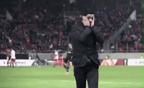 A Man In A Suit Is Standing On A Soccer Field Covering His Face . GIF