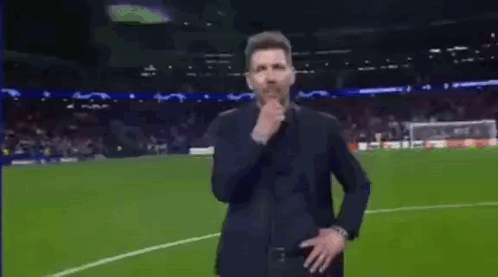 A Man In A Suit Is Standing On A Soccer Field Holding His Hand To His Chin . GIF