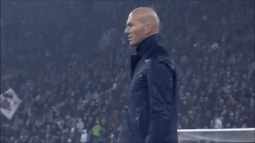A Man In A Suit Is Standing On A Soccer Field Holding His Hand To His Head . GIF