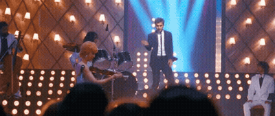 A Man In A Suit Is Standing On A Stage With A Woman Playing A Violin . GIF