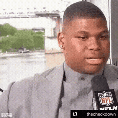 A Man In A Suit Is Talking Into A Microphone While Standing Next To A River . GIF