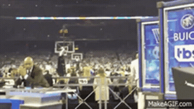 A Man In A Suit Is Talking On A Cell Phone In Front Of A Crowd At A Basketball Game Sponsored By Buick . GIF