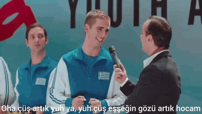 A Man In A Suit Is Talking To A Man In A Blue Jacket With A Greek Flag On His Chest GIF