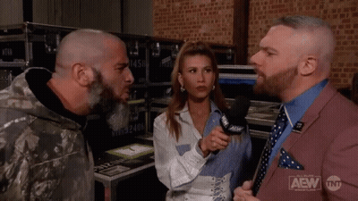 A Man In A Suit Is Talking To A Woman In A Microphone While Another Man Looks On . GIF