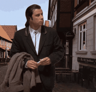 A Man In A Suit Is Walking Down A Street In Front Of A Building With A Sign That Says Kaiser 'S GIF