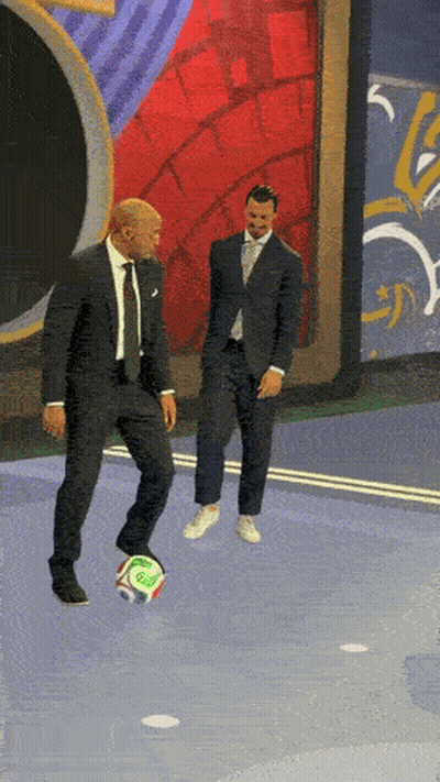 A Man In A Suit Kicks A Soccer Ball While Another Man Stands Behind Him GIF