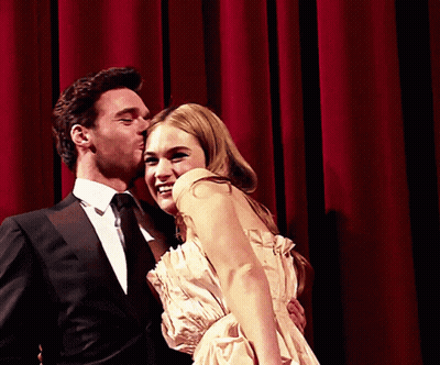 A Man In A Suit Kisses A Woman On The Cheek In Front Of A Red Curtain GIF