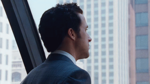 A Man In A Suit Looks Out A Window At The City GIF