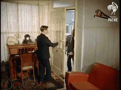 A Man In A Suit Opens A Door Next To A Red Couch GIF