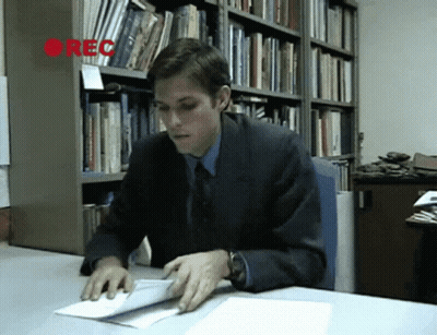 A Man In A Suit Sits At A Desk In Front Of A Bookshelf With The Word Rec On It GIF
