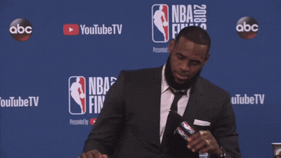 A Man In A Suit Sits At A Table In Front Of A Nba Finals Sign GIF