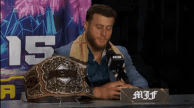 A Man In A Suit Sits At A Table With A Microphone And A Championship Belt On It GIF