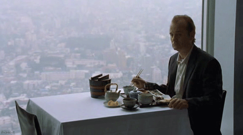 A Man In A Suit Sits At A Table With Chopsticks And A View Of The City GIF