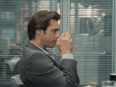 A Man In A Suit Sits At A Table With His Hands Folded In Front Of A Window GIF