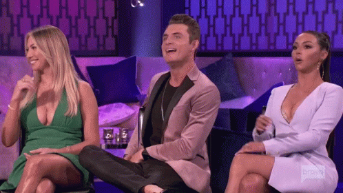 A Man In A Suit Sits Between Two Women On A Couch Laughing GIF
