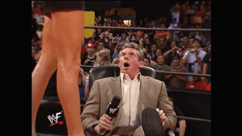 A Man In A Suit Sits In A Chair With A Microphone In Front Of A Wrestling Ring That Says Wwe On It GIF