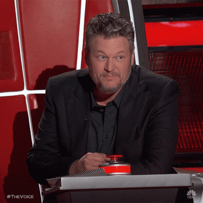 A Man In A Suit Sits In A Chair With A Red Button That Says The Voice On It GIF