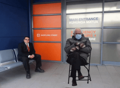 A Man In A Suit Sits Next To A Man In A Chair In Front Of A Sign That Says Main Entrance GIF