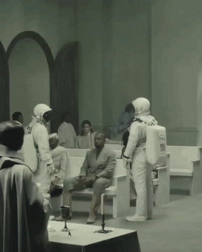 A Man In A Suit Sits On A Bench While A Group Of Astronauts Stand Around Him GIF