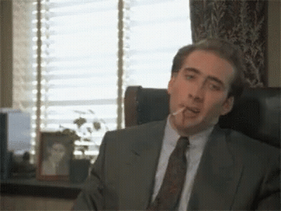 A Man In A Suit Smoking A Cigarette In Front Of A Window GIF