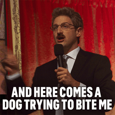 A Man In A Suit Speaking Into A Microphone With The Words And Here Comes A Dog Trying To Bite Me Below Him GIF