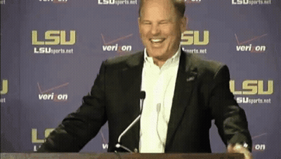 A Man In A Suit Stands At A Podium In Front Of A Wall That Says Lsu On It GIF