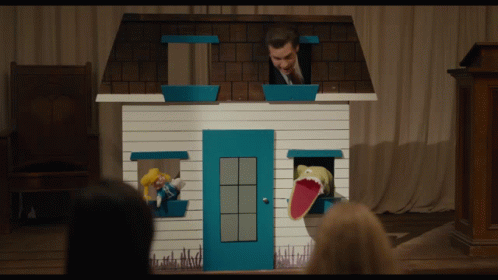 A Man In A Suit Stands Behind A Small House With A Puppet In The Window GIF