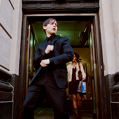 A Man In A Suit Stands In A Doorway While A Woman Walks In The Background GIF