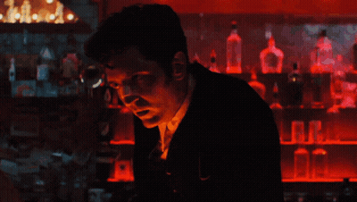 A Man In A Suit Stands In Front Of A Bar With Bottles Of Alcohol On Shelves GIF